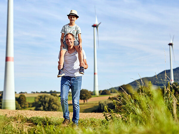 A father carries his son on his shoulders. In the background, wind turbines can be seen.